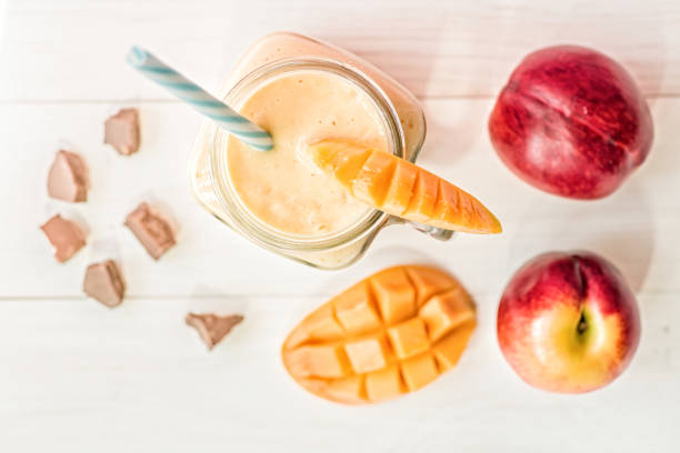 summer drink mango milkshake in mason jar with drinking straw decorated with a half of mango, chocolate pieces flat lay top view on white wooden background