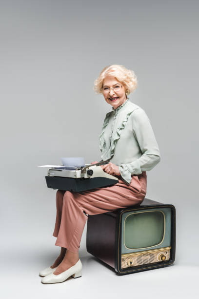istock smiling senior woman using typewriter while sitting on vintage tv on grey 965405224