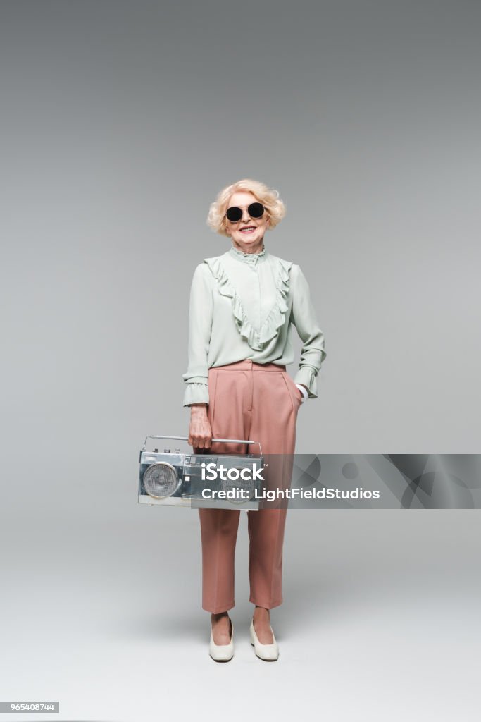 beautiful senior woman in stylish sunglasses with vintage boombox on grey Adult Stock Photo