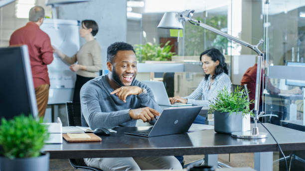istock Man Working at His Desk Working on a Laptop, Encounters Funny Mistake and Laughs. In the Background Creative Young People Doing Their Jobs. Modern Bright Office. 965424420