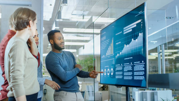 une équipe diversifiée de jeunes professionnels dans la salle de conférence ont discute de statistiques et graphiques montrés sur un téléviseur de présentation. - television commercial photos et images de collection