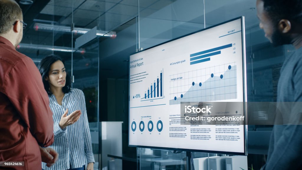Beautiful Hispanic Woman Analyzes Statistics, Charts and Pies with Company's Growth Shown on a Wall TV. Presentation - Speech Stock Photo