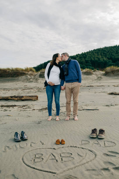 Mum, dad and baby written on the sand with the parents kissing behind Mum, dad and baby written on the sand of the beach next to their shoes with the parents kissing behind funny pregnancy announcements stock pictures, royalty-free photos & images