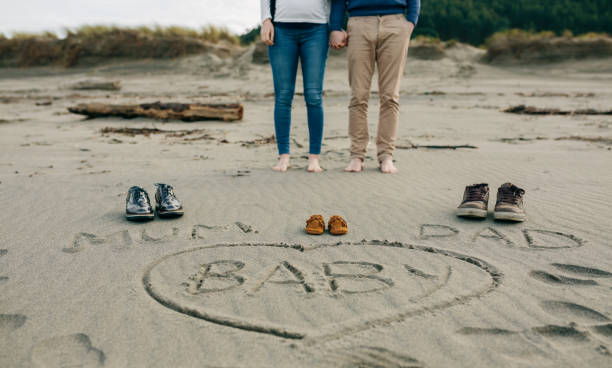 Mum, dad and baby written on the sand with the parents behind Mum, dad and baby written on the sand of the beach next to their shoes with the legs of the parents behind funny pregnancy announcements stock pictures, royalty-free photos & images