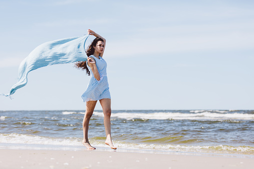 Pretty girl walking by the sea with a blue waving scarf. Summer by the sea.