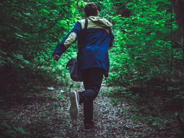 young scary man running away in the dark forest on the path back view