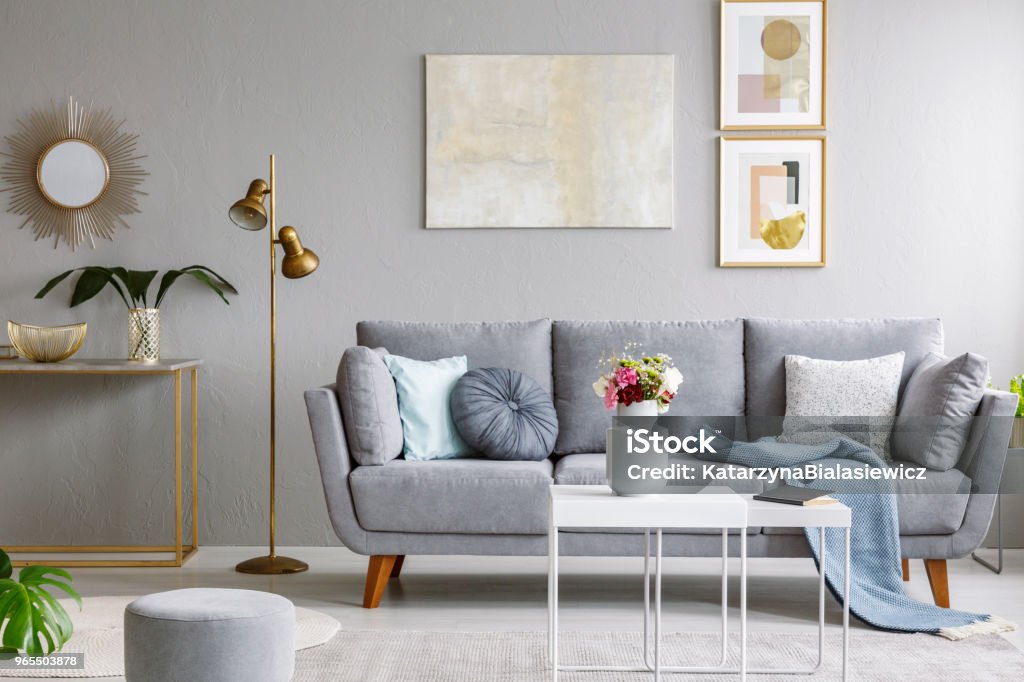 Gold mirror above shelf with plant in grey living room interior with sofa and flowers on table. Real photo Gold mirror above shelf with plant in grey living room interior grey sofa and flowers on table Living Room Stock Photo