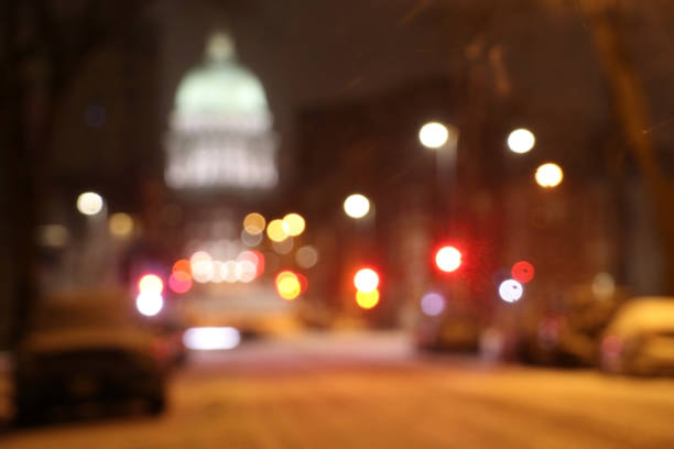 Downtown street view with silhouette go glowing in the dark State Capitol building and parked cars during snowy winter night. Madison, Wisconsin, Midwest USA.