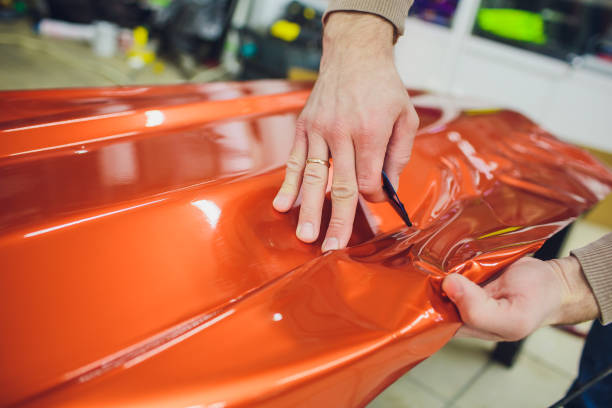 Car wrapping specialist putting vinyl foil or film car wrapping protective film yacht, boat, ship, car, mobile home. orange film heating with hair dryer and trimming plastic soft hard squeegee stock photo