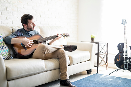 Portrait of handsome young hispanic man sitting on sofa and playing guitar