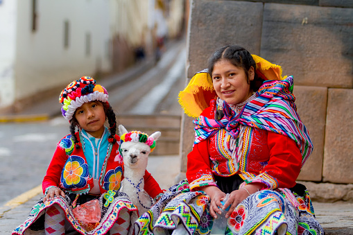 Cusco, Peru - February 10, 2018: Young woman with her little niece pose in a portrait with their typical costumes next to her little alpaca in a street in the historic center of Cusco