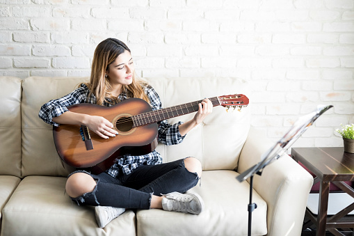 Beautiful 20 years old woman sitting on couch and playing guitar in her living room