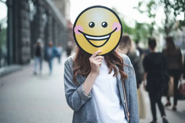 Cheer up. Waist up portrait of lady is standing in the street with yellow laughing emoticon. She is replacing her face with smiley
