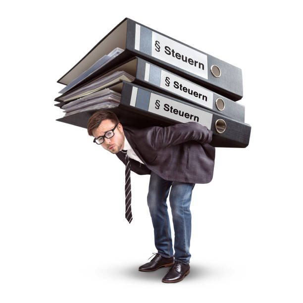 A businessman with a straining expression is carrying a large stack of folders. The folders have the German word \
