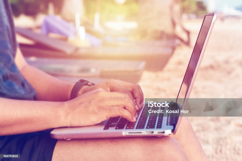 A man wearing shorts with laptop working on a summer day on the beach A man wearing shorts with laptop working on a summer day on the beach. Studying, Working, Technology, Freelance Work Concept. Laptop Stock Photo