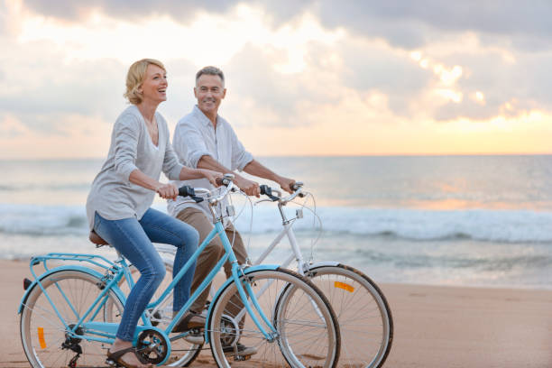 coppia matura in bicicletta sulla spiaggia al tramonto o all'alba. - mature adult bicycle senior adult heterosexual couple foto e immagini stock