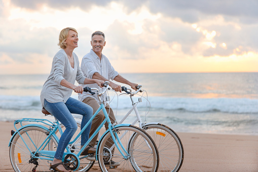 Mature couple cycling on the beach at sunset or sunrise. They are laughing and having fun. They are casually dressed. Could be a retirement vacation.