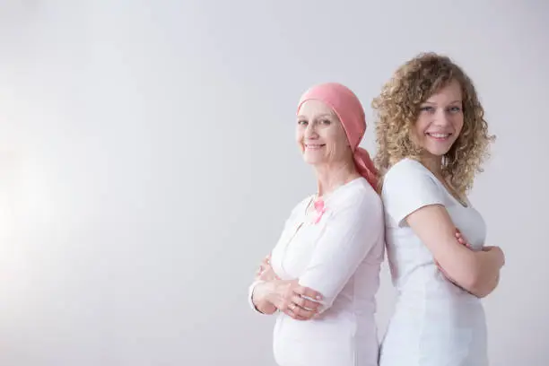 Female cancer patient with a pink headscarf and ribbon and her daughter standing happy back to back on a gray empty background