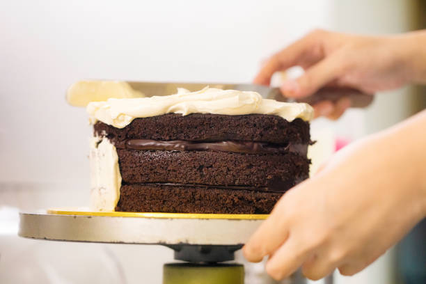 Close-up on baker's hands placing butter cream icing on layered Chocolate cake using a spatula