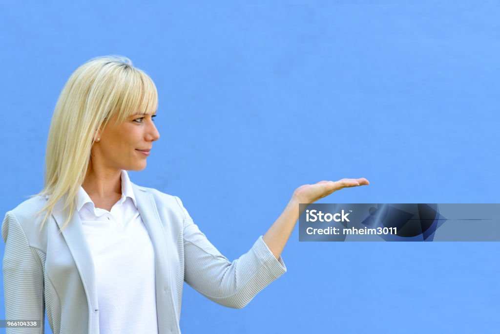 Smiling attractive blond woman holding out a hand Smiling attractive blond woman holding out her empty hand for product placement over blank copy space over a yellow studio background Adult Stock Photo