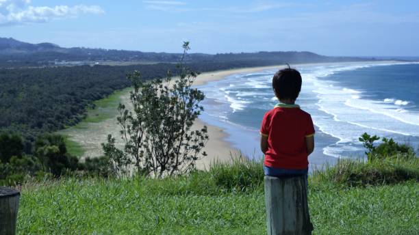Kid sitting on lumber post looking at panoramic view of ocean, land. Shot from behind child sitting on timber post enjoying nature landscape of sea, forest, mountains. Lonely kid or thinking kid concept.