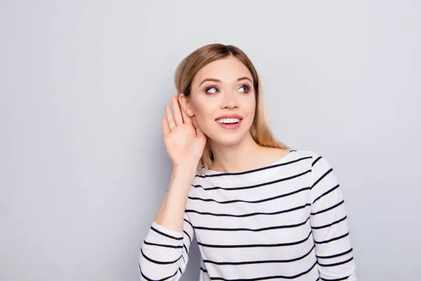 Portrait of charming, pretty, stylish, trendy, nice, cute woman in striped sweater with cupped hand curiously listening to gossip or secret over gray background