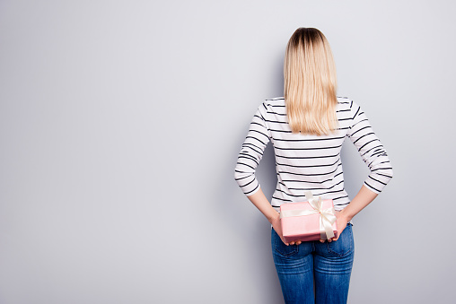 Rear view portrait with copy space for advertisement of slim, pretty girl having gift in pink package with white bow in arms, prepare surprise, isolated on grey background