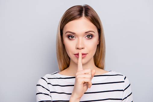 Shhh! Close up portrait of charming, pretty, stylish woman in striped outfit with hairstyle showing silence symbol, holding forefinger on lips, looking at camera, isolated on grey background