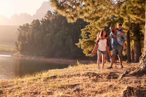 parents giving children piggyback rides on walk by lake - two parent family couple family african ethnicity imagens e fotografias de stock