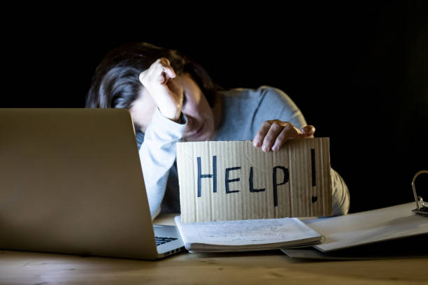 istock Young tired and stressed student at home working late at night on her laptop holding a help sign in a over worked and addicted concept on isolated black background 966224714