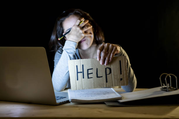 istock Young tired and stressed student at home working late at night on her laptop holding a help sign in a over worked and addicted concept on isolated black background 966224718