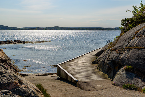 Back lit bathing bridge and ramp into the sea on a sunny morning. Suns reflection in the water. The planks follow the natural rock to the right. Hovik on the island Tjorn, Sweden.