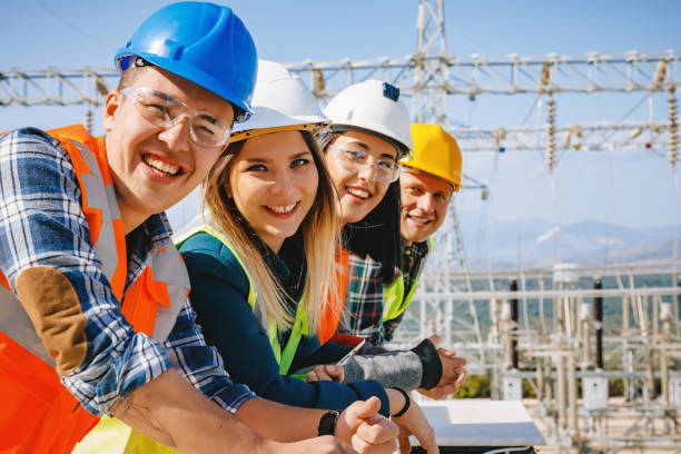 Group portrait of young workers in electric power station Group of young men and women supervisor business people, electrician, engineer and technician workers working and look at camera posing in front of electrical components and towers of high tension of an electricity power generation station field in a sunny day rural landscape. xxxl size electrician smiling stock pictures, royalty-free photos & images