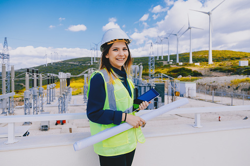 Portrait of engineer worker businesswoman in white hard-hat and safety vest holding blueprints working and look at camera posing on by wind turbine power generation station in rural landscape