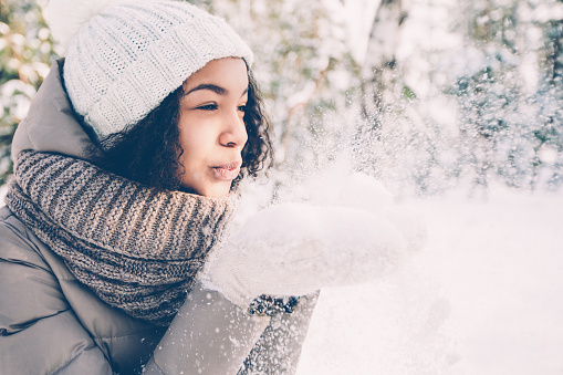 Portrait of happy teenage Latin American girl wearing white knit hat, scarf and jacket blowing snow off her hands in winter