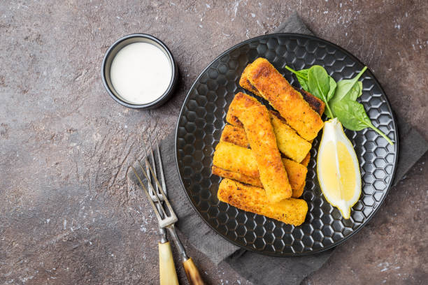 Crumbed fish fingers Tasty savory snack of crumbed fish fingers sticks served on a plate with lemon over dark stone background, top view with copy space fish stick stock pictures, royalty-free photos & images
