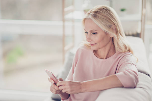 nonna affascinante, carina, elegante e moderna seduta sul divano in salotto a casa, facendo shopping online attraverso internet 3g, avendo lo smartphone in mano - furniture internet adult blond hair foto e immagini stock