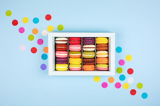 Box of colorful macarons on blue background with confetti. Flat lay style.
