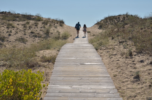 Two unrecognizable persons walk away on the wooden footpath going through sandy dunes to sea beach. Bulgaria. Slightly blurred image.
