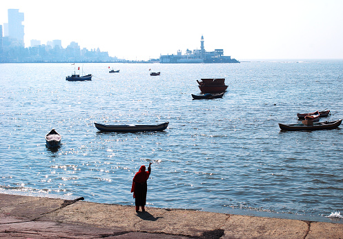 A woman devotee offering flowers to Arabian Sea and praying in Mumbai with country fishing crafts bobbing on the waves. Skyscrapers and other structures of south Mumbai can be seen in the background. Copy-space
