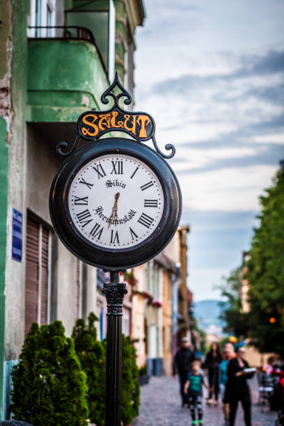 Close up color image depicting an old fashioned clock outdoors on the ancient, medieval street of Sibiu, a city in the Transylvania region of Romania. On the clock face is the name of the city (Sibiu) and also its German name - Hermannstadt. At the top of the clock is the word 'Salut', which is a welcoming greeting in Romanian. Beyond the clock, out focus, people are walking on the streets of Sibiu. Room for copy space.