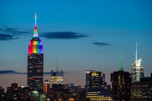 A partial skyline of Midtown Manhattan with the Empire State Building displaying Pride colors taken from Brooklyn just before at sunset.