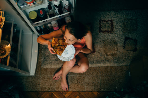 woman eating in front of the refrigerator in the kitchen late night - one slice imagens e fotografias de stock