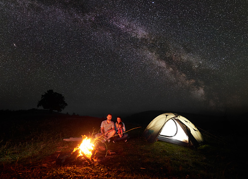 Modern tourist family camping in mountains by night. Husband and wife with small daughter sit on tree trunk in front of tent at burning campfire and look in camera. Tourism and traveling concept.
