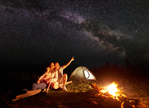 Tourist family camping in mountains at night, sitting on log near illuminated tent and burning bonfire. Mother holding in arms small daughter, father pointing at bright stars in dark sky and Milky way