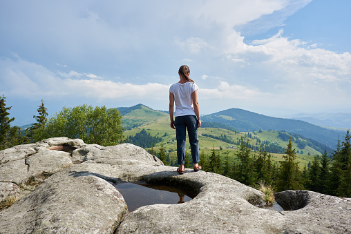 Back view of young long-haired tourist girl standing on huge rock with big puddle in the middle under beautiful blue sky enjoying fantastic view of mountains covered with ever-green pine forest.