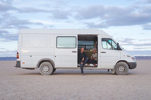 An adorable mixed-race senior couple sit in the open doorway of their white camper van and make music together. She is playing the ukulele. They are parked in the dry desert flats with nothing around except a mountain range in the distant background.