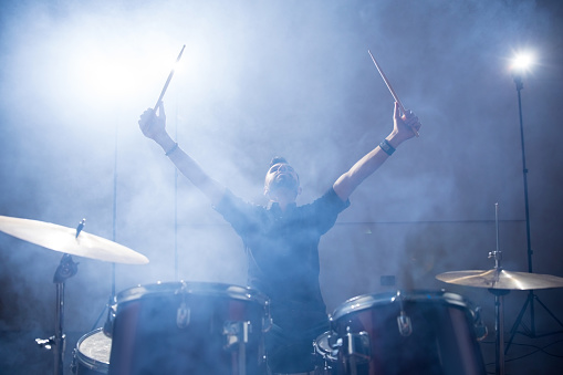 Rock band drummer raising his arms on stage with light and smoke