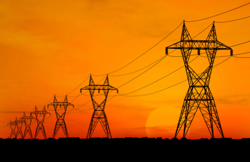 Several electric power lines rising above a black foreground.  A bright orange sky is seen in the background during sunset.