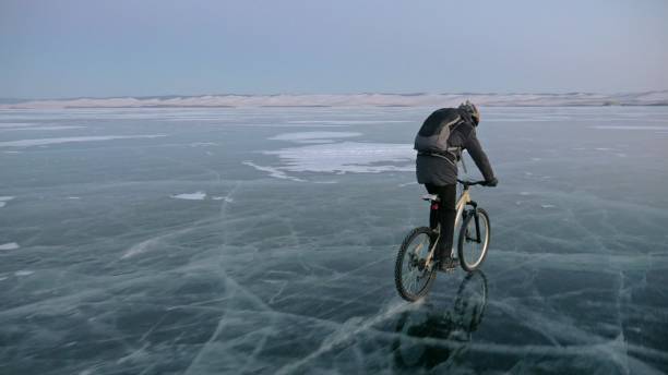 istock Man is riding a bicycle on ice. The cyclist is dressed in a gray down jacket, backpack and helmet. Ice of the frozen Lake Baikal. The tires on the bicycle are covered with special spikes. 966750326